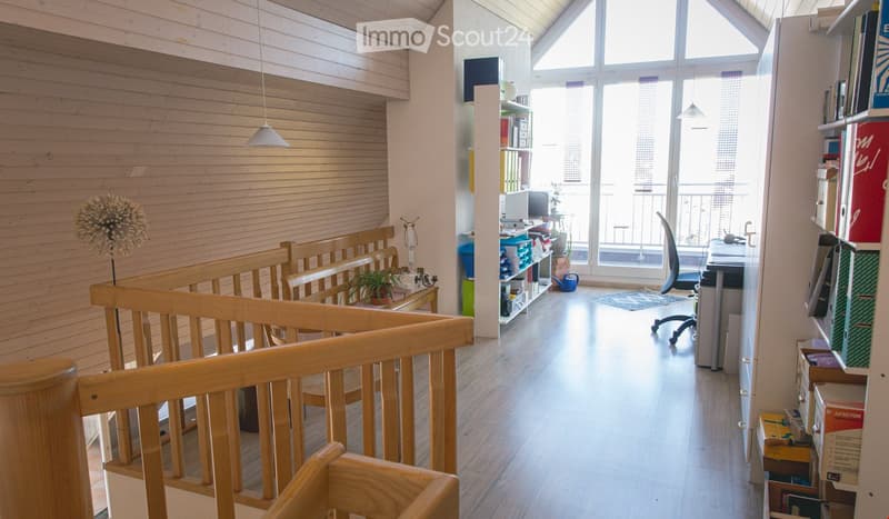 The image shows a well-lit, modern hallway with wooden flooring and white walls. There is a wooden railing on the left side, leading to a seating area with a small table and chairs. On the right side, there is a desk with a computer, shelves filled with b