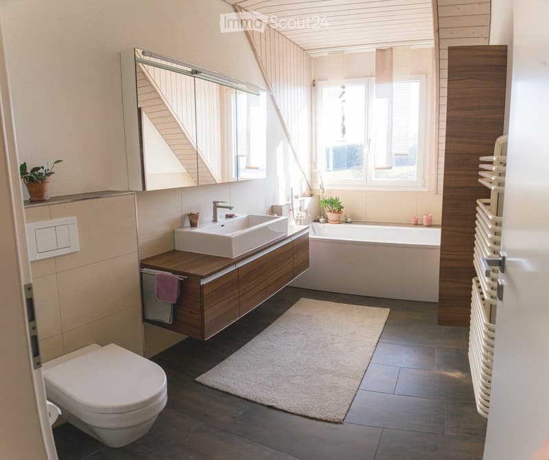 Modern bathroom with a large sink, toilet, bathtub, and a large mirror. Features a wooden bathroom cabinet, a white rug, and a radiator on the wall.