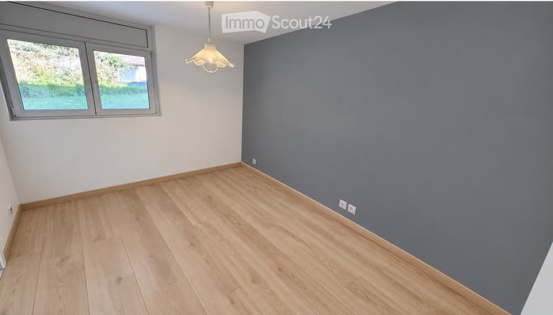 Empty room with wooden flooring, grey and white walls, and a hanging light fixture. The room has two windows with a view of greenery outside.