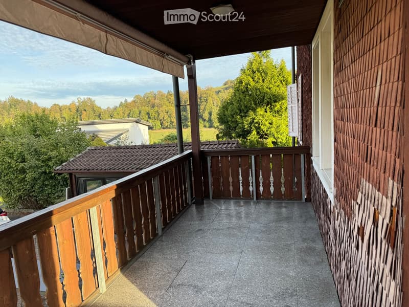 Balcony with wooden railing, tiled floor, and scenic view of trees and a house in the background.