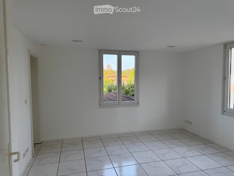 Empty room with white walls, tiled floor, and a window with a view of the roof and trees outside.