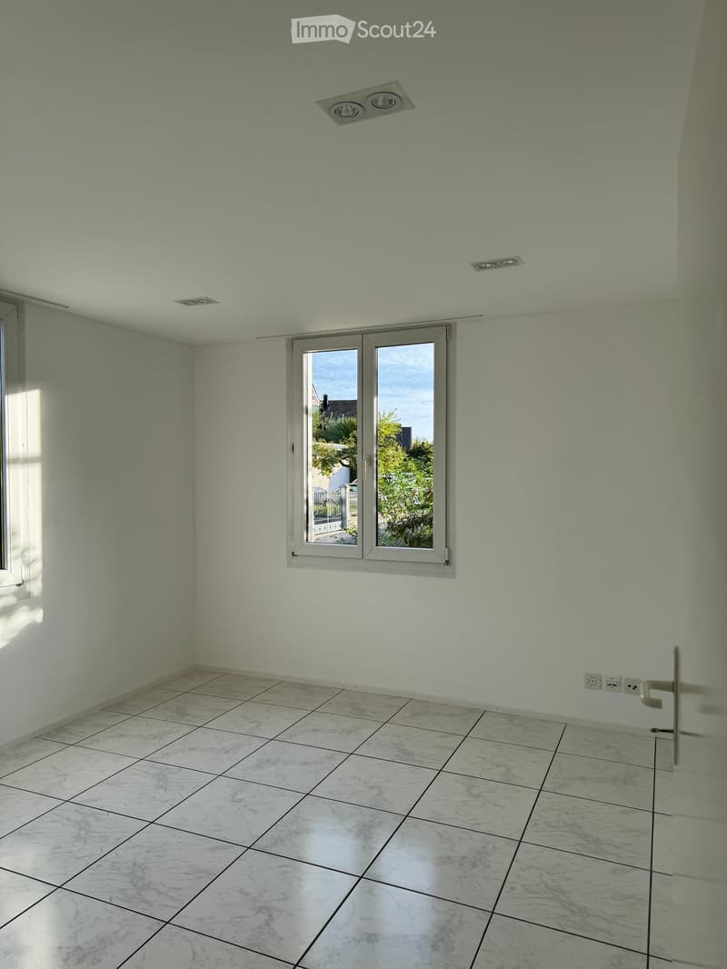 Empty room with light green walls, marble tiled floor, and a large window with a view of the outside. The room has recessed ceiling lights and electrical outlets on the wall.