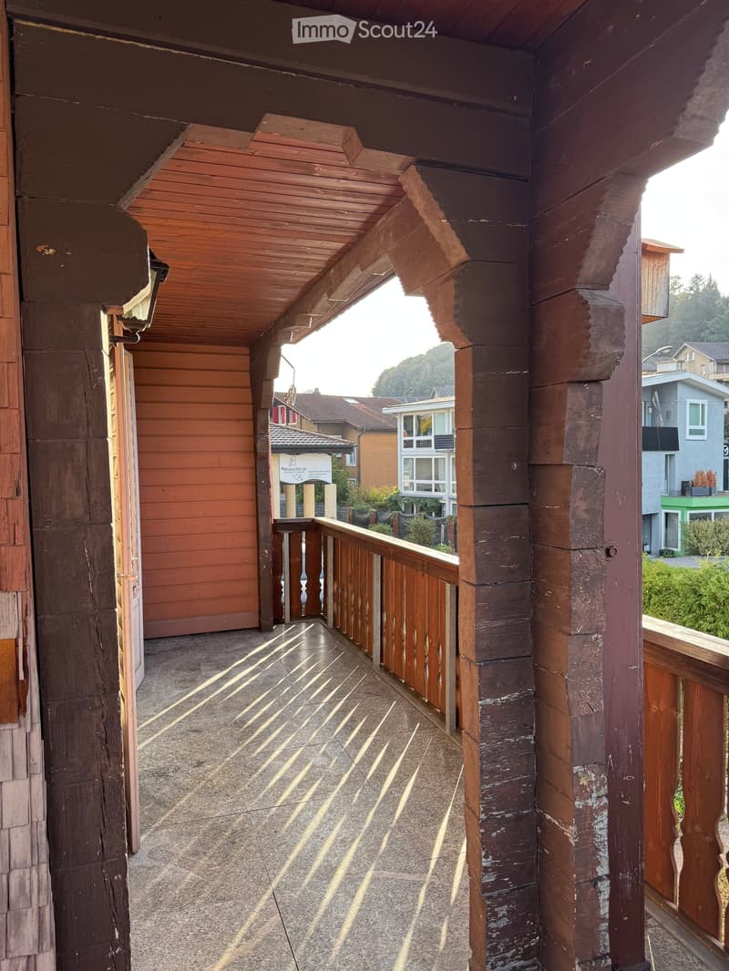 Wooden balcony with a covered roof, wooden railings, and a view of neighboring houses and greenery.