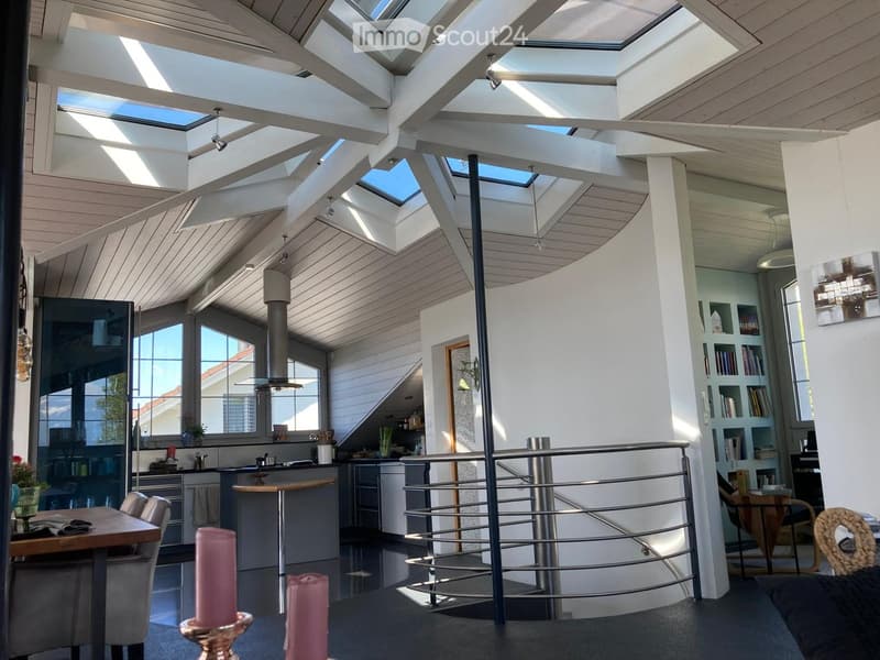 Bright, open-plan living and dining area with a high, slanted wooden ceiling and skylights. Features a modern kitchen island, dining table with chairs, bookshelf, and decorative candles.