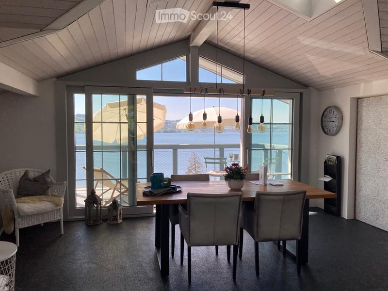 Bright dining area with large windows offering a scenic view, featuring a wooden dining table, chairs, a wicker chair, and decorative items.
