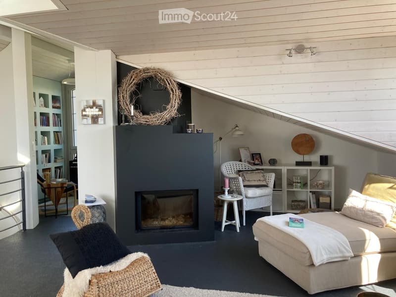 Modern living room with a fireplace, sofa, chairs, a small table, and a bookshelf. The room features a dark grey fireplace with a decorative wreath on top, a white wicker chair, and a beige sofa with pillows.