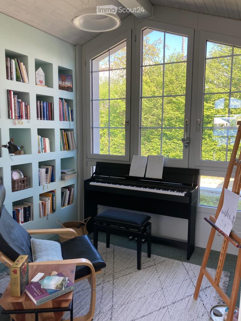 Well-lit room with large windows, a black piano with a bench, a wooden easel, a comfortable chair, a small table with books, and built-in bookshelves.