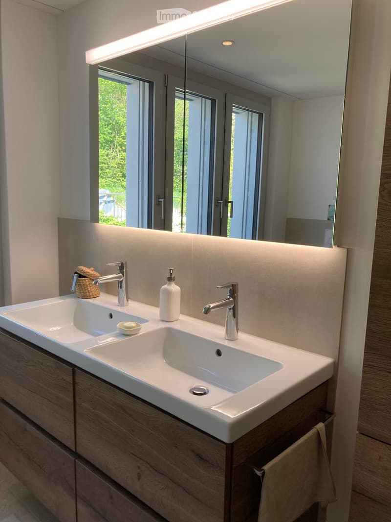 Modern bathroom with a double sink, sleek chrome faucets, a large mirror with integrated lighting, and a wicker basket on the counter.
