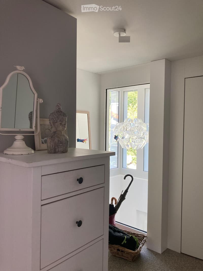 A small, well-lit hallway with a dresser, mirror, lamp, and a decorative statue on top. An umbrella and a wicker basket are placed near the door leading to a balcony.