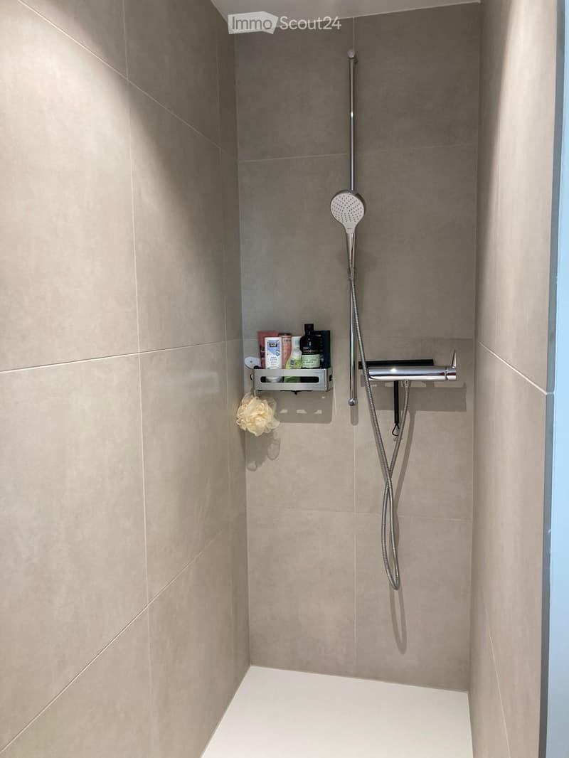 Modern shower area with beige tiles, a handheld showerhead, a shower shelf holding various products, and a decorative flower hanging from the shelf.