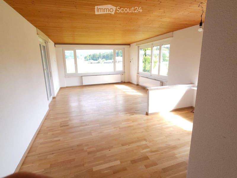 Empty room with light wooden flooring, white walls, large windows, and a wooden slanted ceiling.