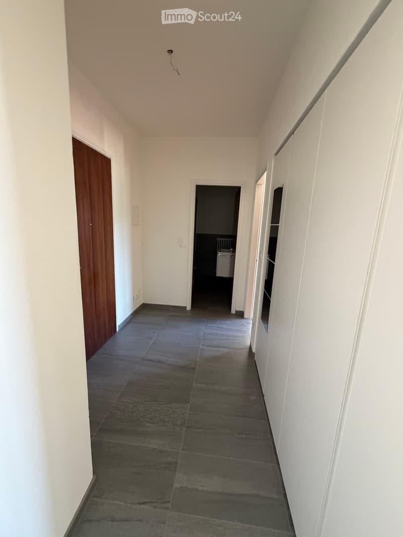 Empty hallway with tiled floor, white walls, and wooden door on the left. A partially visible room with a radiator is seen at the end.