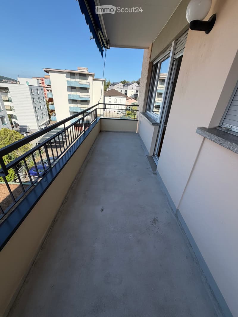 Empty balcony with a metal railing, overlooking a residential area with buildings and parked cars.