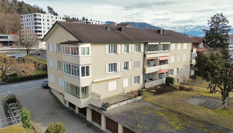 Multi-story residential building with beige facade, multiple windows, balconies, and surrounding greenery with mountain view.