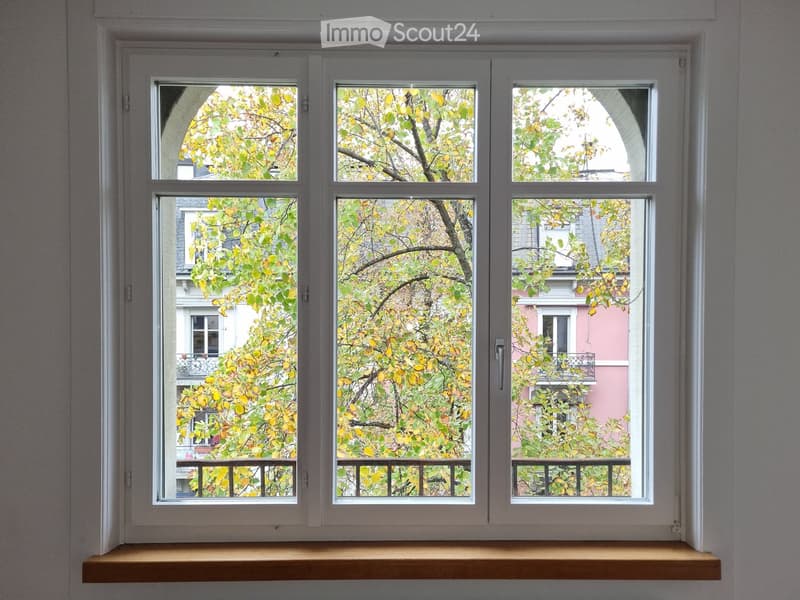 An unfurnished room with a large window offering a view of autumn foliage and neighboring buildings. The window has multiple panes and is framed in white, with a wooden windowsill below.
