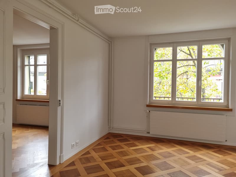 Empty room with light-colored walls, wooden flooring, and large windows showing greenery outside. Radiators are visible under the windows.