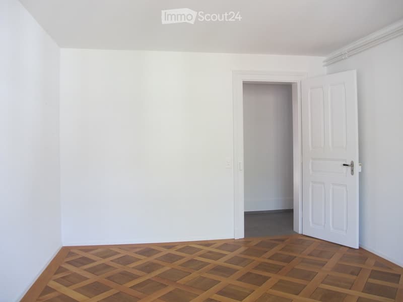 Empty room with white walls, wooden floor, and an open door leading to another room.