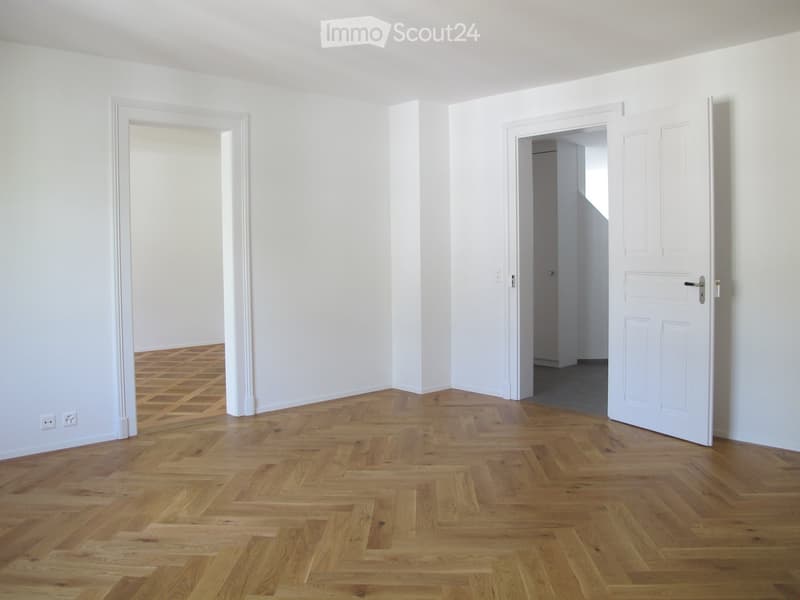 Empty room with white walls and herringbone wooden flooring, featuring two open doorways leading to adjacent spaces.