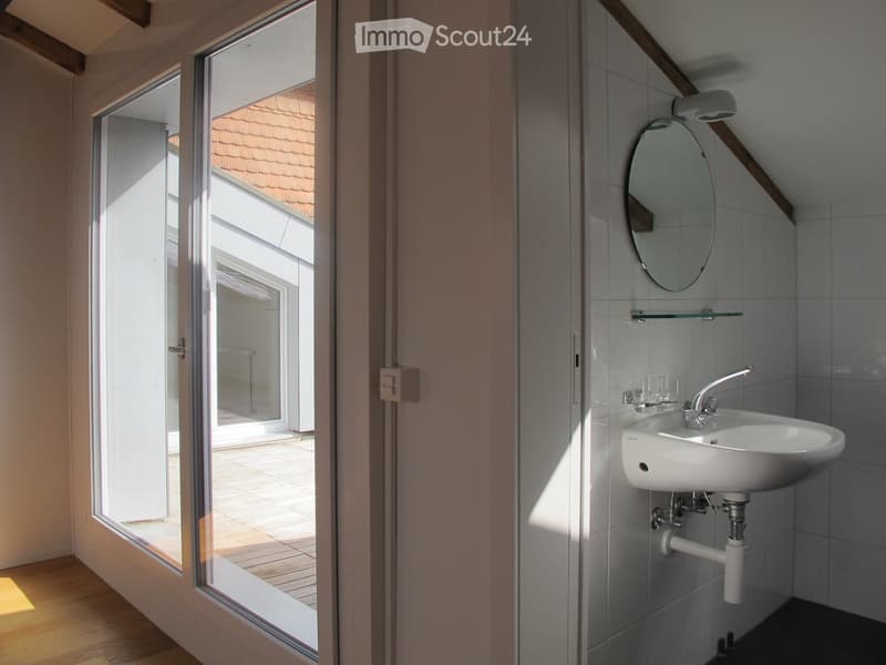 Modern bathroom with a white sink, chrome faucet, round mirror, and glass shelf. Large windows provide natural light and a view of the wooden deck outside.