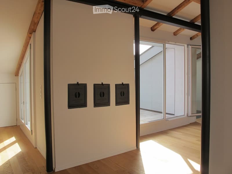 Bright hallway with wooden flooring, exposed wooden beams, and large windows allowing natural light to flood the space. Three black wall-mounted devices are visible on the wall.