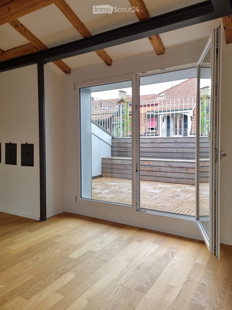 Bright, empty room with wooden flooring and exposed wooden beams on the ceiling. Large sliding glass doors open to a wooden deck and outdoor staircase leading to a rooftop terrace.