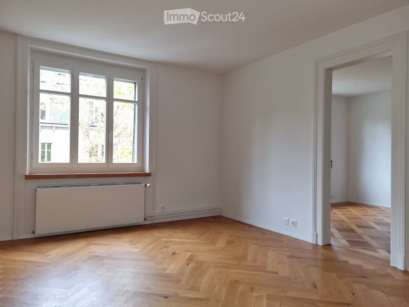 Empty room with wooden flooring, white walls, large windows, radiator, and a doorway leading to another room.
