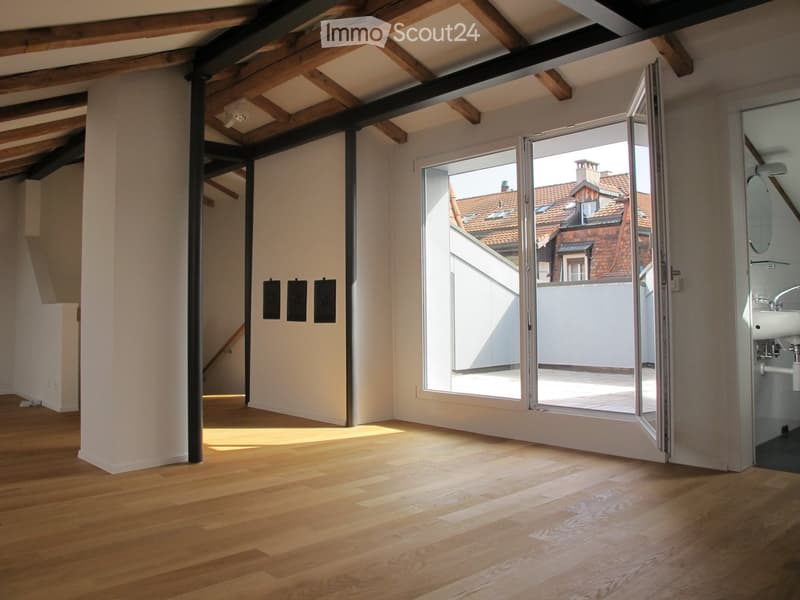 Empty room with wooden flooring, exposed wooden beams on the ceiling, large sliding glass doors leading to a balcony, and a small adjacent bathroom area visible.