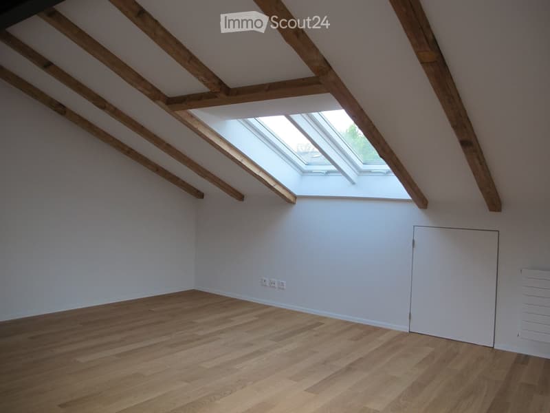 Empty attic room with wooden beams, skylight window, wooden floor, and white walls.