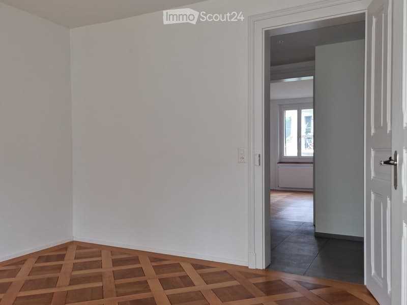 Empty hallway with white walls and wooden flooring, leading to another room with a window and radiator.