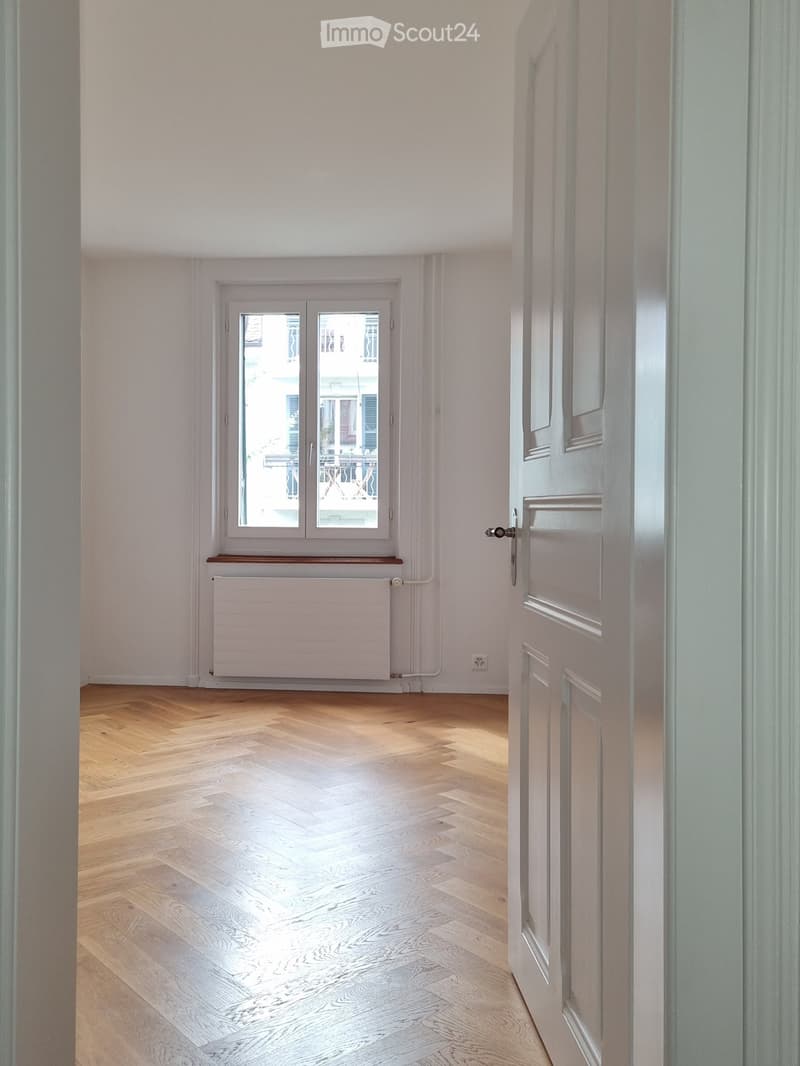 Empty room with wooden flooring, white walls, a window with double glass panes, a radiator, and a closed door.