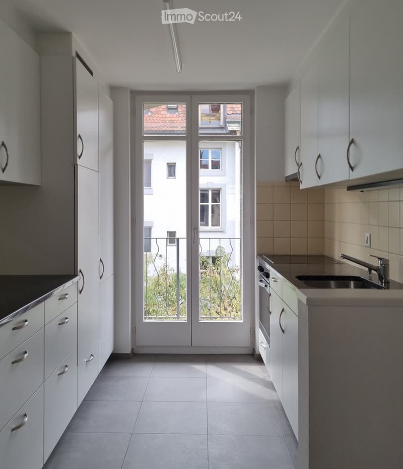 Modern kitchen with white cabinets, a sink, and an oven, featuring large windows and a door leading to a balcony.
