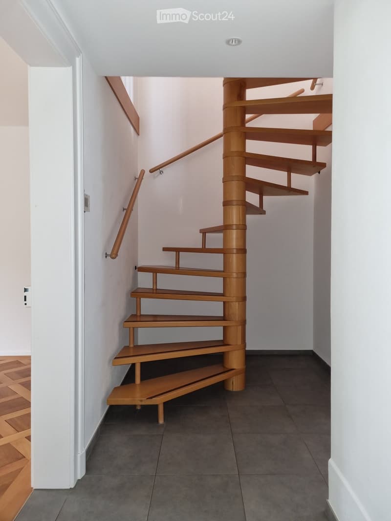 Spiral wooden staircase with open wooden railings, white walls, and polished wooden floor in the foreground.