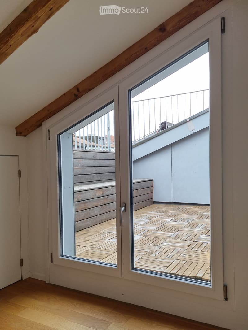 View from inside a room showing two large windows opening to a wooden decked balcony area with white railings and wooden steps leading up.