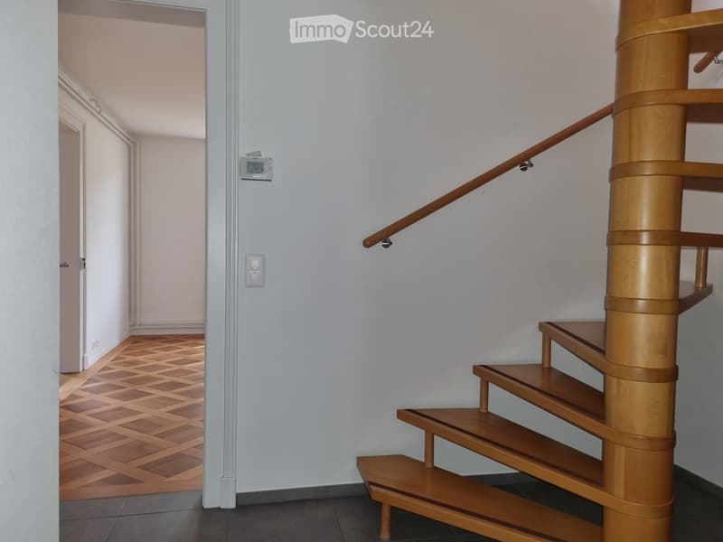 Spiral staircase with wooden steps and handrail, adjacent hallway with patterned wooden flooring and white walls.