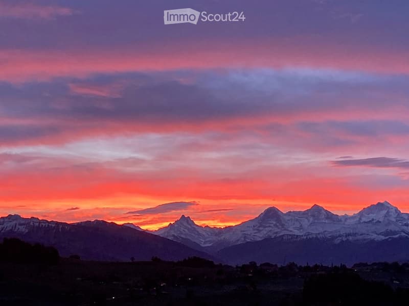 A scenic view of snow-capped mountains at sunset, with a vibrant sky of pink and orange hues, and a small settlement visible in the foreground.