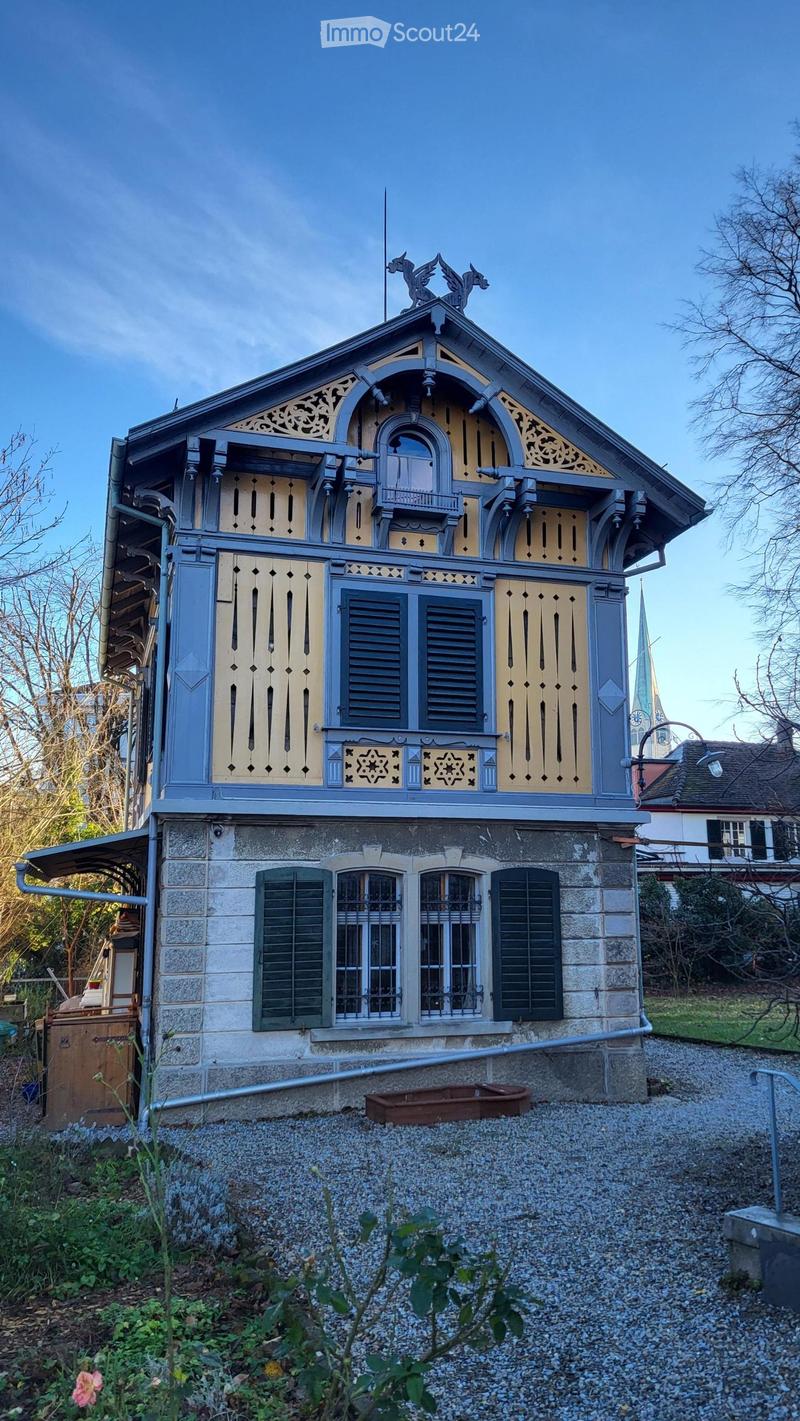 Two-story house with ornate wooden detailing, blue and cream exterior, stone base, multiple windows with shutters, decorative roofline, and small balcony.