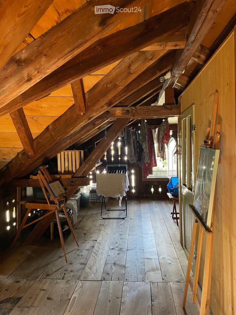 An attic space with exposed wooden beams and wooden flooring, featuring a chair, stool, table, mirror, and clothing rack.