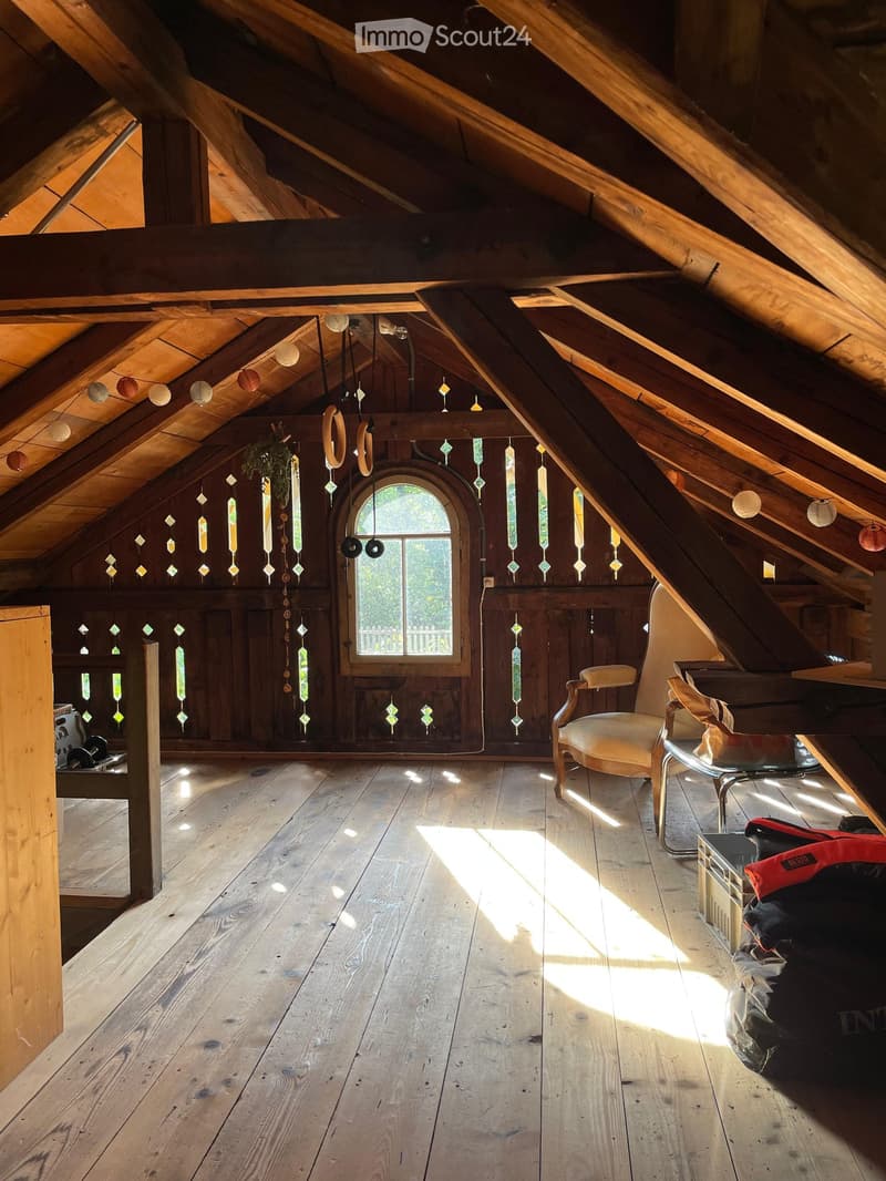 Cozy attic space with wooden floors, slanted wooden ceiling beams, and colorful stained glass windows. Features a small seating area with a chair, table, and stool.