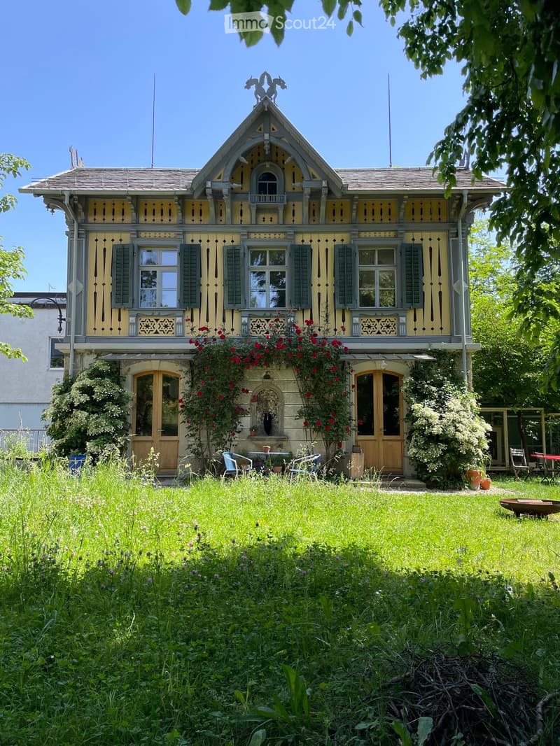 Two-story house with yellow and grey exterior, decorative wooden trim, multiple windows, and a well-maintained garden with outdoor furniture.