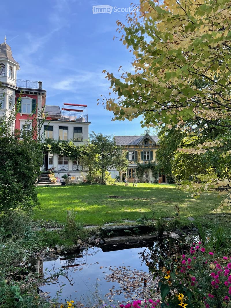 A lush garden with a pond, surrounded by trees and flowers, with several historic houses in the background.