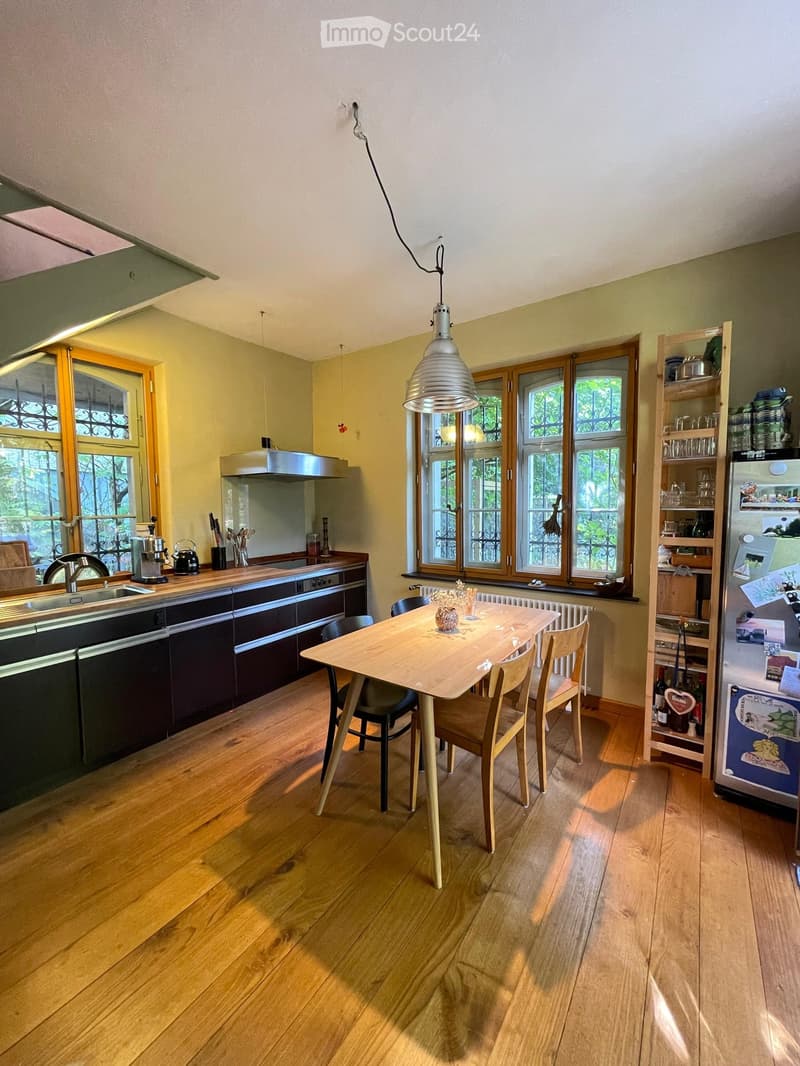 Bright kitchen with wooden flooring, large windows, and modern appliances. Includes a sink, coffee maker, teapot, knife block, dishwasher, range hood, dining table with chairs, and a refrigerator with shelves.