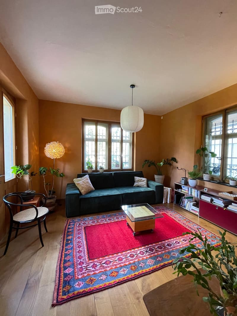 Living room with wooden flooring, large windows, a dark green sofa with throw pillows, a glass coffee table, a colorful rug, a wicker chair, a bookshelf, and several potted plants.