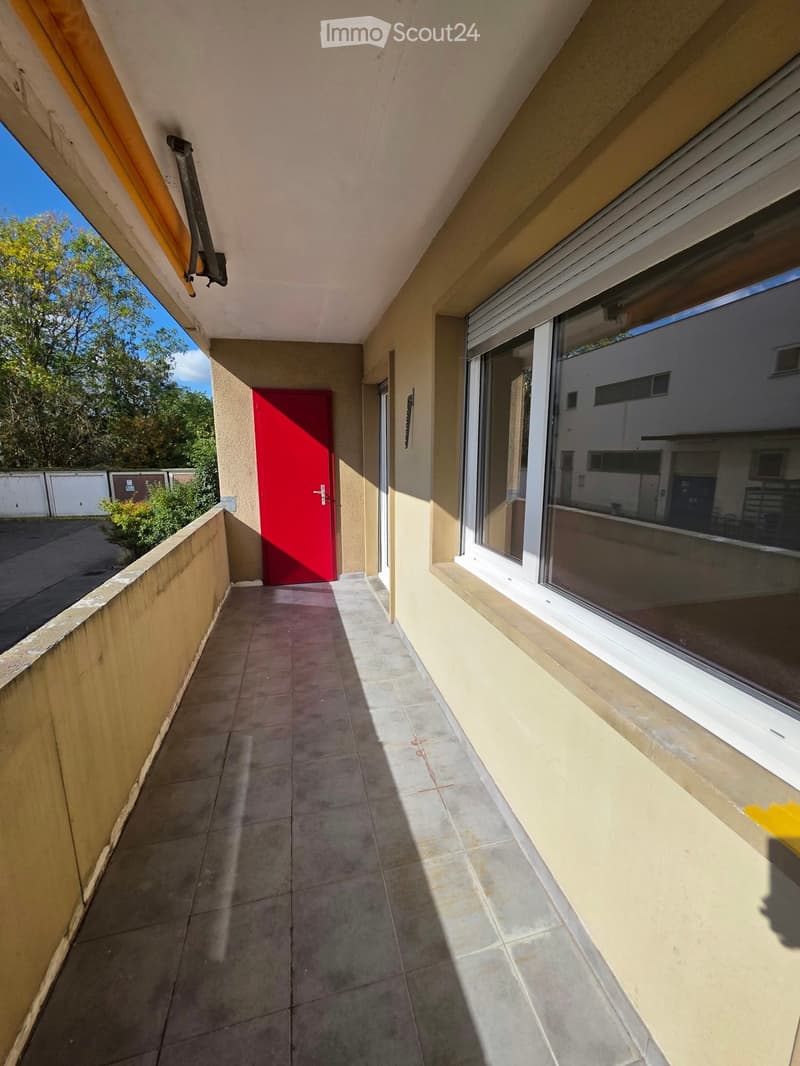 Balcony with tiled floor, beige walls, large window, red door, and view of greenery and parking area.