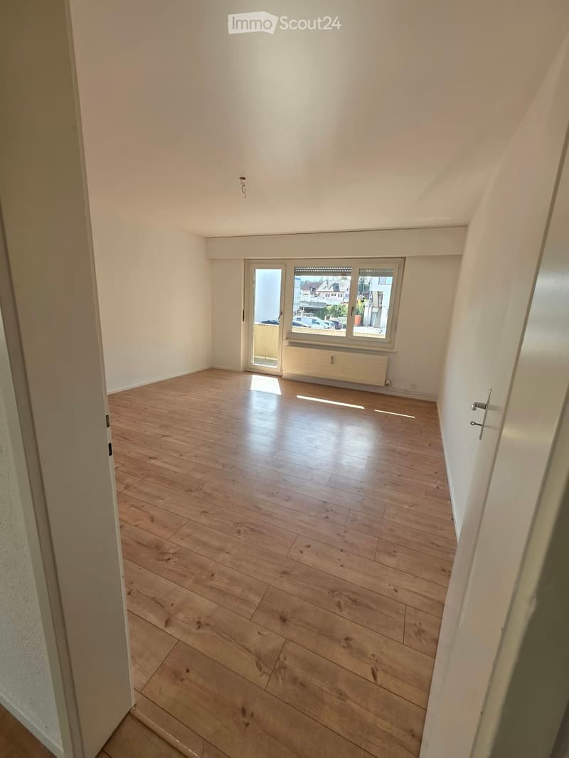 Empty room with wooden flooring, white walls, and large windows offering a view of the street and parked cars.