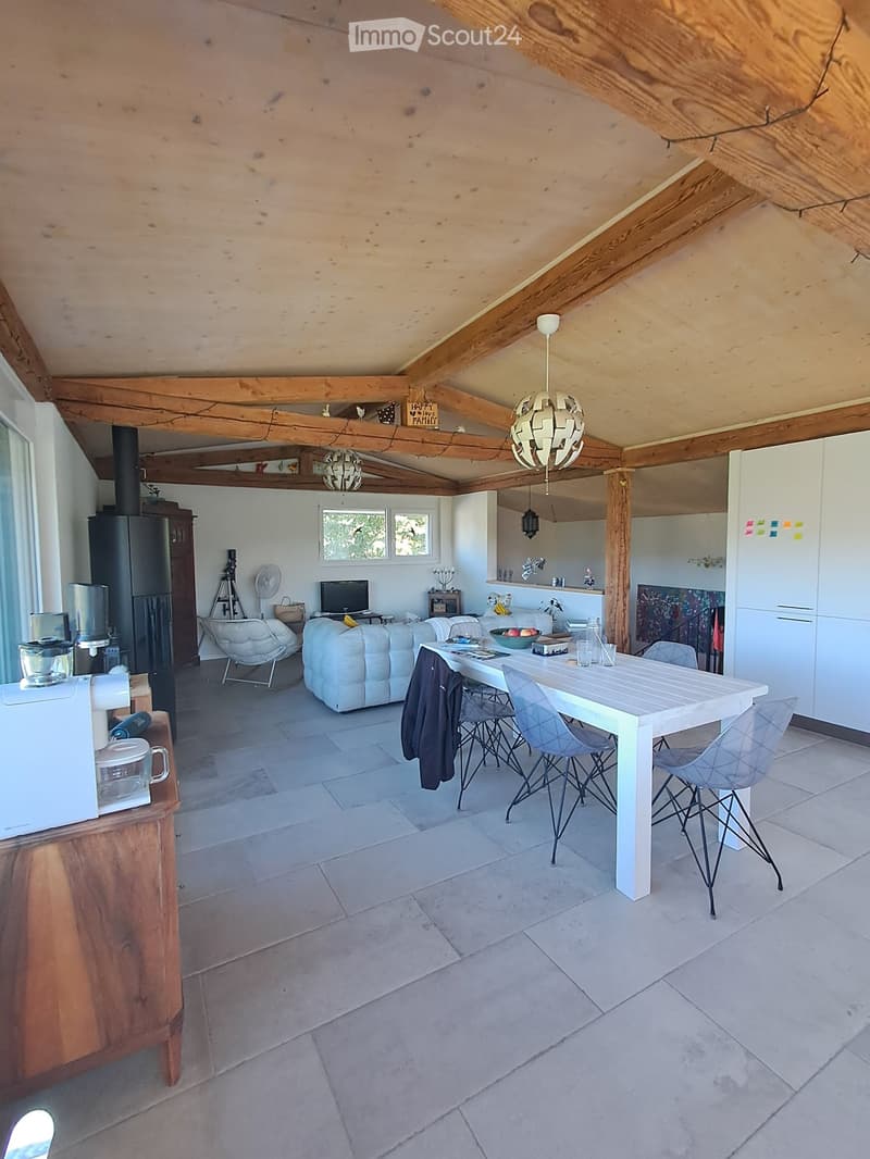Open-plan living and dining area with exposed wooden beams, a white sofa, a dining table with chairs, a refrigerator, and a television. The space features a tiled floor and modern lighting.