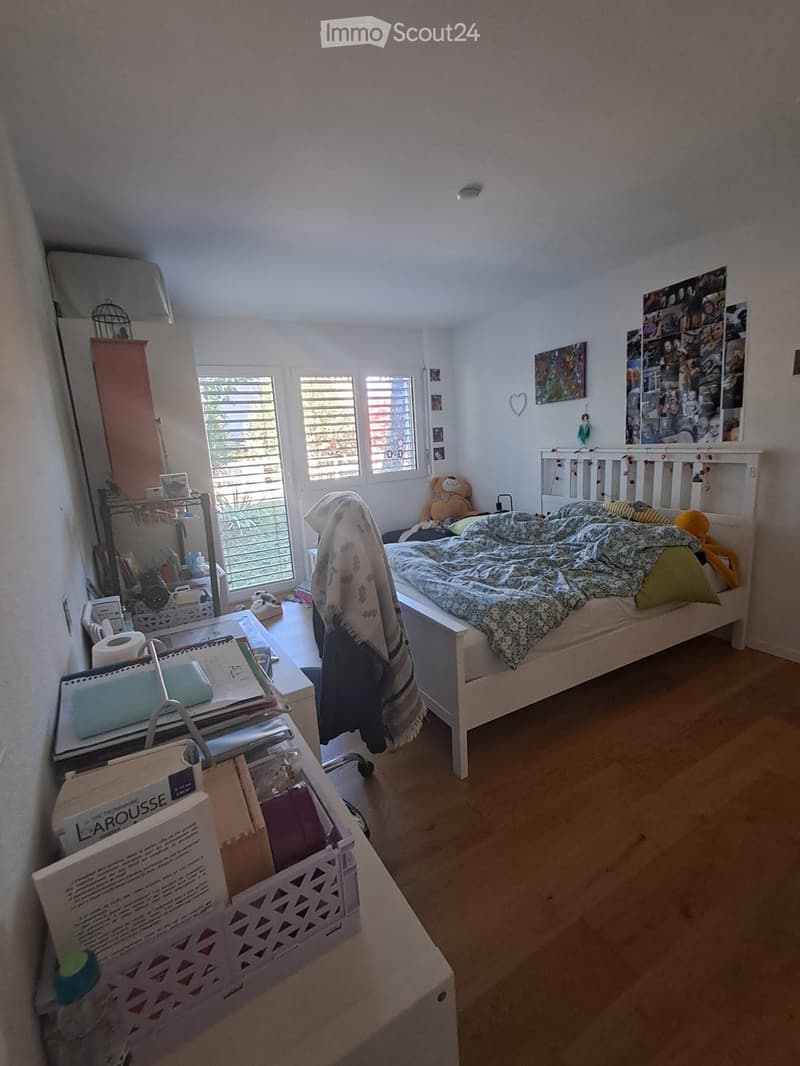 Cozy bedroom with a single bed, bedside table, mirror, chair, and storage basket. The room features wooden flooring and large windows with blinds, allowing natural light to fill the space.