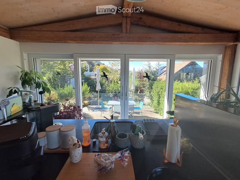 Modern kitchen with wooden beams, large windows overlooking a garden, sink, coffee maker, and various kitchen items on the counter.