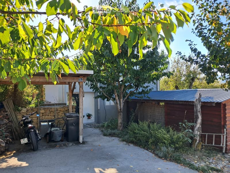 Outdoor area with a covered wooden pergola, a motorcycle, a small shed, and various greenery including trees and shrubs.