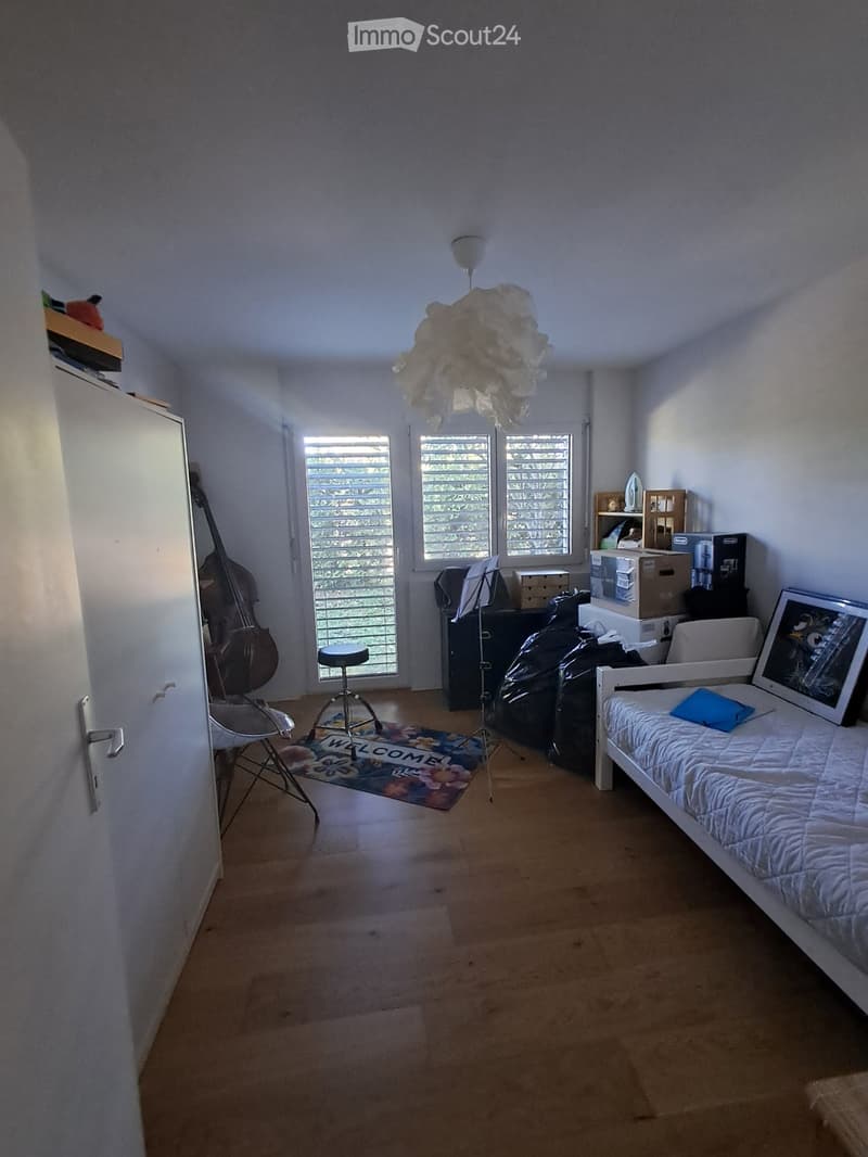 Bright bedroom with wooden flooring, large windows, bed with white bedding, a chair, a stool, a cello, a picture frame, and a lamp.