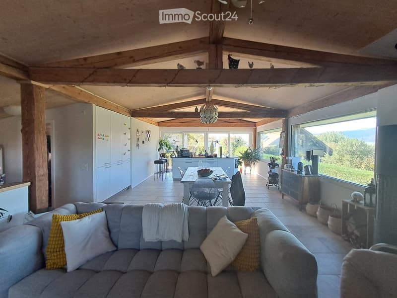 Open-plan living and dining area with exposed wooden beams, a grey sectional sofa with yellow and white cushions, a wooden dining table with chairs, a modern kitchen with white cabinets and stainless steel appliances, and large windows offering a scenic v